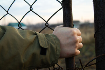 Hand in a Khaki Sleeve Grasping a Rusted Metal Fence Post with Wire Mesh