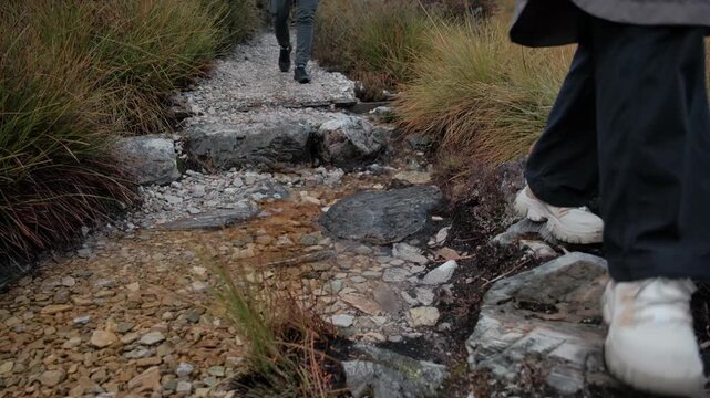 Close-up of hikers' feet stepping across a clear stream on a rocky trail. Trekking through alpine wilderness in Cradle Mountain National Park, Tasmania.