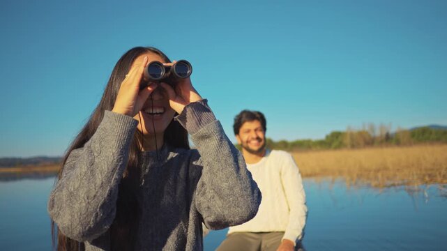 A young woman uses binoculars by a serene lake, while a man smiles in the background. The scene captures exploration and connection, ideal for outdoor activities, couple relationships.