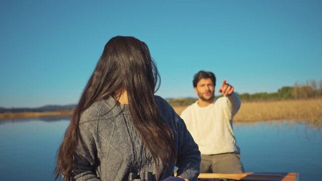 A couple enjoys a serene day outdoors by a calm lake. The woman uses binoculars while the man points into the distance, embodying a relaxed, active lifestyle and fostering connection with nature.