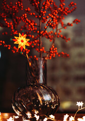 Elegant Ilex berries bouquet in a glass vase with a glowing Christmas star and warm fairy lights....