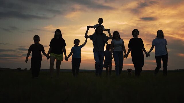 Walking together at sunset silhouetted family cross open field with father carrying child while other child holds hand with parent under sky and horizon creating silhouette and sunset mood warm