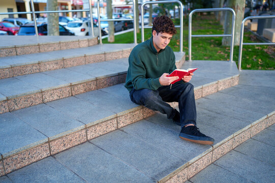 Young man reading red book, sitting on concrete steps outdoors, focusing on education and casual learning