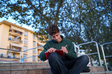 Young man studying outdoors, sitting on urban steps and concentrating while writing or drawing in a red notebook