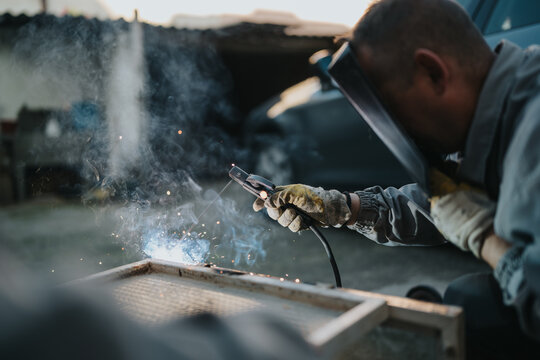 Skilled welder wearing protective gloves and a mask uses a welding torch to fuse metal, producing bright sparks in an industrial workshop. Focus on craftsmanship, safety, and industrial fabrication.
