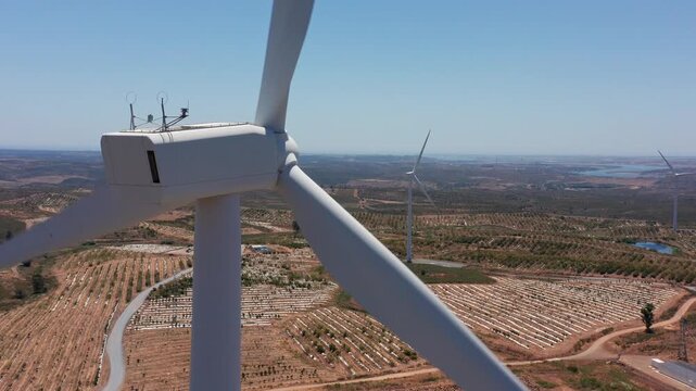 aerial wind turbine close-up in spain showing rotating blades and nacelle above arid farmland, winding service road, distant turbines on horizon, clear blue sky, drone inspection perspective