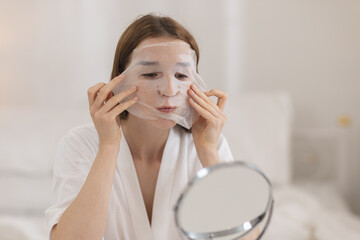 women wearing white bathrobe adjusting sheet mask on her face while looking into mirror representing skincare routine hydration and beauty treatment with self care.
