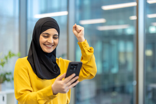 Muslim woman wearing a hijab and yellow shirt triumphing with a raised fist while looking at her smartphone in a modern office with blurred background, representing digital success and achievement - Powered by Adobe