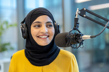 Young muslim woman wearing a hijab and headphones. Smiling while actively engaging in a podcast or radio broadcast. Communicating with an audience through a professional microphone in a studio setting