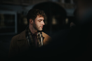 A stylish man wearing a brown coat and plaid scarf stands in a dim outdoor setting, captured with moody lighting that emphasizes fashion and introspective emotion.