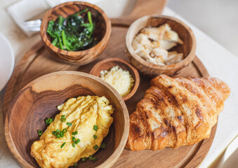 A wooden platter holds a variety of breakfast foods, including a croissant, scrambled eggs, and sautéed greens. The presentation is complete with wooden bowls showcasing Parmesan and another side.