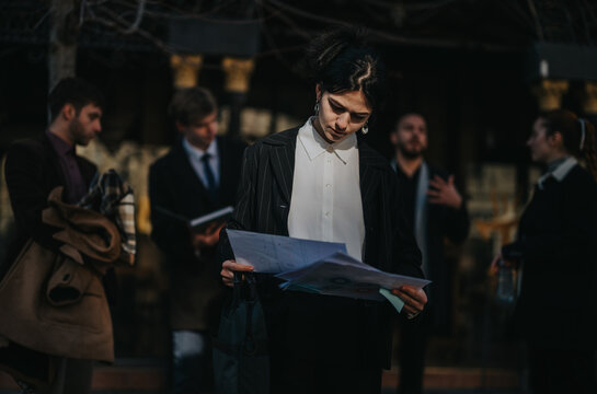 A businesswoman in a pinstriped suit examines papers as a group of colleagues mingle in the background, conveying corporate work, collaboration, and decision making in an urban office environment.