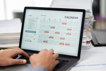 A hand checks a laptop calendar to manage a deadline timeline with an overworked task planner in...