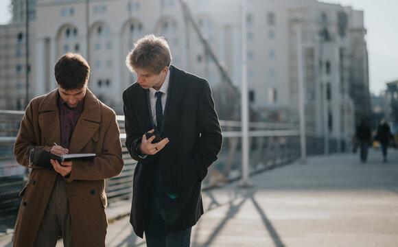 Two young colleagues walk along a modern urban path, one taking notes while the other checks a smartphone. Dressed in smart coats, they project a professional, collaborative vibe in a city setting.