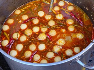 Overhead close-up of a metal pot holding Jungle Curry Noodles, or Khanom Chin Nam Ya Pa, with fish balls simmering in a spicy dark-orange broth with red chilies and herbs. A popular, spicy Thai dish.