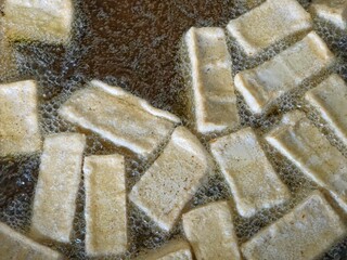 Close-up overhead shot of multiple rectangular pieces of tofu being deep-fried in bubbling hot oil.