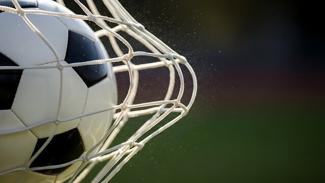 Dynamic close-up of a classic black-and-white soccer ball striking the net with visible motion, capturing the decisive moment of a goal on a bright outdoor football field
