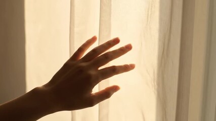 A delicate hand reaches toward a window, gently touching sheer curtains backlit by soft, golden morning sunlight, evoking warmth and privacy. - Powered by Adobe