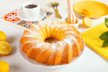A freshly baked lemon bundt cake dusted with powdered sugar, served on a white plate with sliced lemons, a cup of coffee, and vibrant tableware on a cozy, bright background.