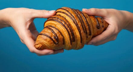 Two hands cradle a chocolate-drizzled croissant against a vibrant blue backdrop