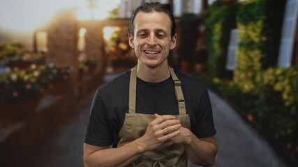 Man with long hair wearing apron, hands clasped on a street with potted plants and blurred storefronts; warm welcome.