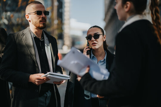 A small group of business colleagues stands on a city street, reviewing documents while one person talks on a mobile phone. They wear coats and sunglasses, conveying a busy outdoor meeting.