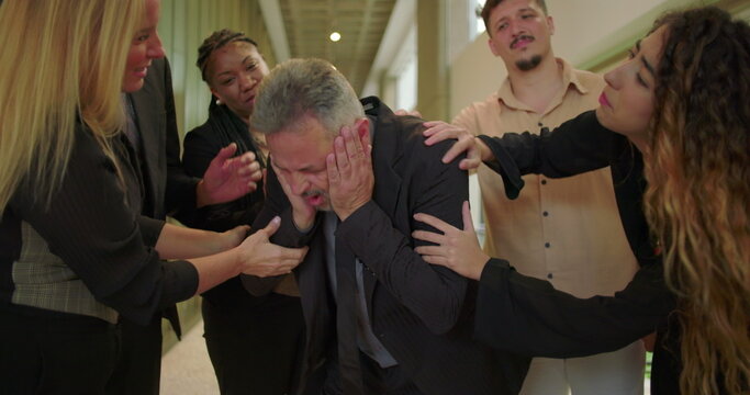 Distressed male professional showing anguish and worry while being comforted by supportive colleagues in office hallway during emotional workplace moment