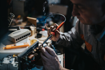 A technician uses a soldering iron to repair an electronic device at a cluttered workbench, with tools and components nearby, capturing hands-on craft and precision in a busy workshop.