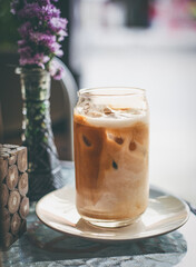 An iced coffee sits on a plate, bathed in soft light. A vase with purple flowers adds a delicate touch to the setting.