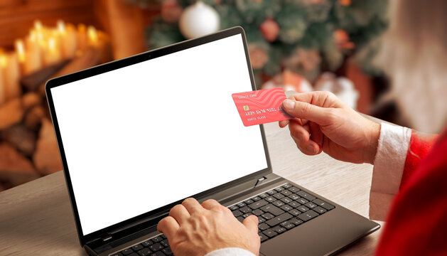 Santa holding credit card while using laptop with blank screen in warm Christmas interior, ideal for holiday mockups, ecommerce concepts and festive digital marketing