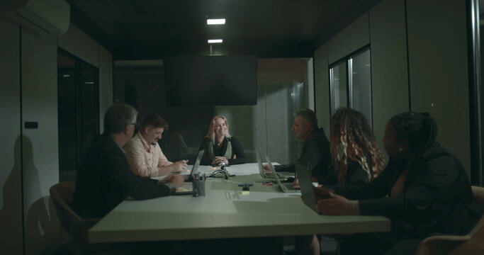 Diverse business team in serious discussion around boardroom table during meeting, using laptops and tablets, seated across modern office conference room - Powered by Adobe