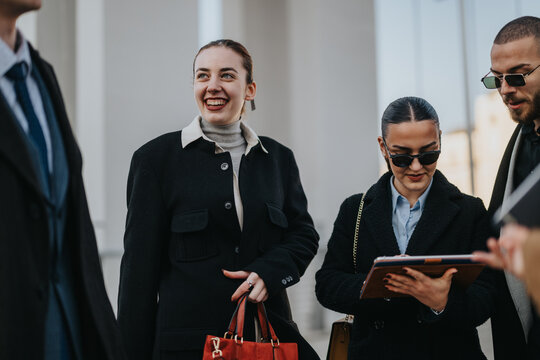 A group of young coworkers stands outside, smiling and chatting as one holds a tablet and a red bag. Modern, confident colleagues enjoying a casual business moment. - Powered by Adobe