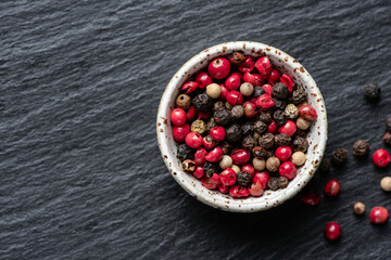 Group of red black and white peppercorns in white ceramic bowl on black slate, top view copy space