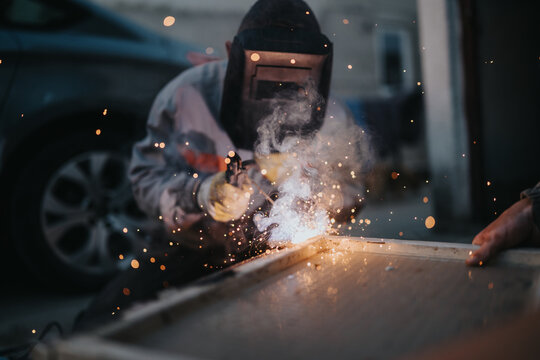 A skilled welder in full protective gear works on a metal surface, producing vivid sparks and smoke. The scene highlights industrial labor, safety equipment, and the energy of fabrication.