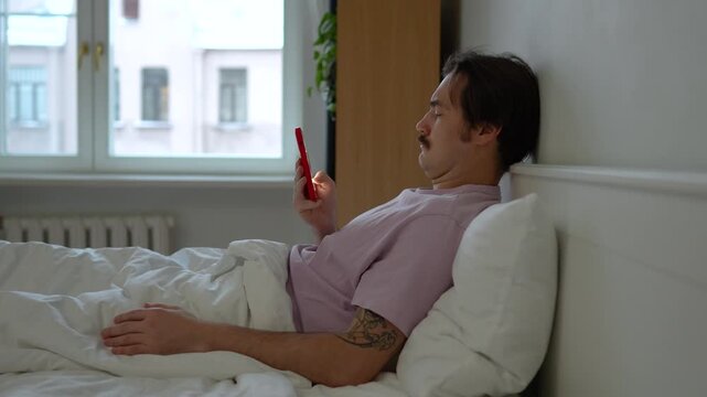 Morning with social media. Procrastinated man in using smartphone gets glued to phone, getting quick dopamine from scrolling. Interested male sits under blanket in bed checking phone after wake up.