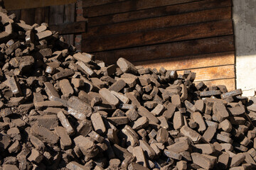 A large pile of dark peat briquettes is seen in a storage area, resting against a rustic wooden wall