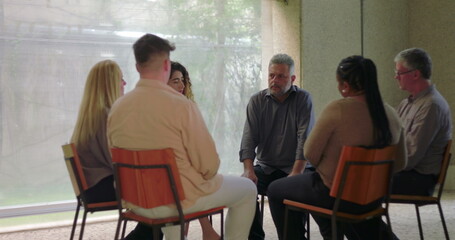 Diverse group of adults sit in circle during emotional group therapy session, engaging in serious conversation in quiet, supportive indoor setting with natural light
