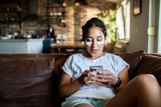 Young adult woman relaxed using smartphone with eye patches at home