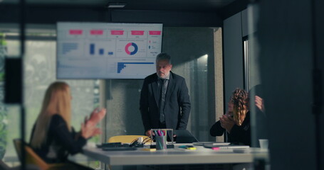 Diverse team applauding business leader following successful data presentation in modern corporate boardroom with screen displaying charts