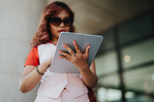 A stylish woman in pink business casual attire stands outdoors, using a tablet. Sunglasses and accessories complete her modern, professional look as she focuses on digital tasks in a sunny setting.