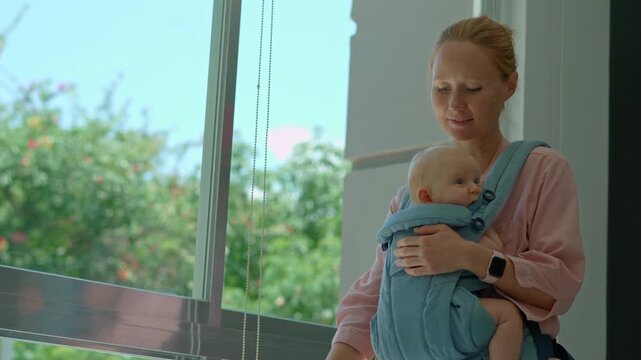 Young mother washes the floor in a bright apartment with a large window and lush greenery outside, while her newborn baby sits safely in a front ergonomic baby carrier