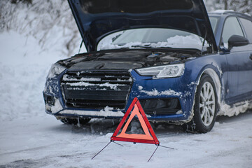 Blue car with open hood and warning triangle on a snowy winter roadside