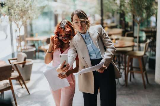 Two women in stylish business attire pose for a selfie while reviewing papers in a bright cafe setting. Their teamwork and casual energy highlight collaboration and everyday business moments.