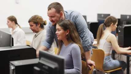 In classroom during computer courses, male teacher helps girl student complete difficult task, looks at monitor screen and corrects work. Mentor comments on students work, gives hints during task