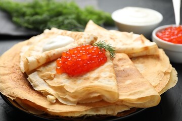 Delicious crepes with red caviar and sour cream on black table, closeup