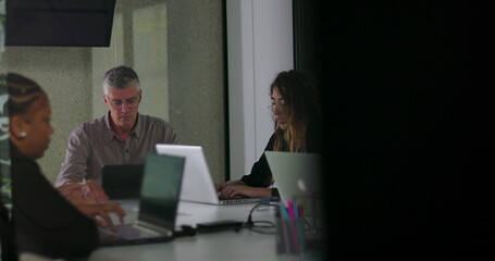 Focused business team working on laptops during quiet office meeting with diverse colleagues seated around table in modern workspace