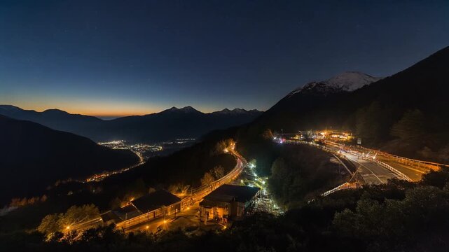 Snowy mountain valley at dusk with winding road and glowing buildings beneath gradient sky