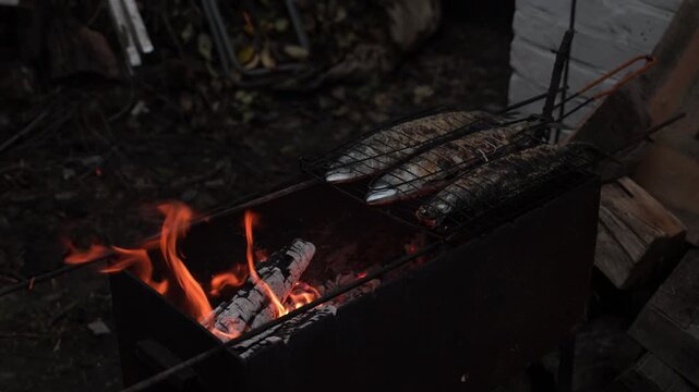 Three mackerel are grilled over an open fire, forming a golden-brown crust. The fish rest on the grill, enveloped in smoke.