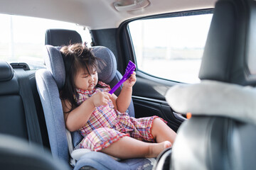 A cute little Asian girl is sitting in a child car seat inside a car and is happily playing with toys.