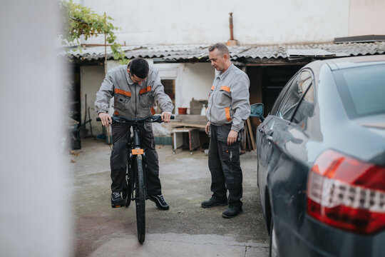Two technicians in gray uniforms examine a bicycle while a car sits nearby in a yard. They appear focused on maintenance, showcasing teamwork and practical mechanical skill in an outdoor workspace. - Powered by Adobe